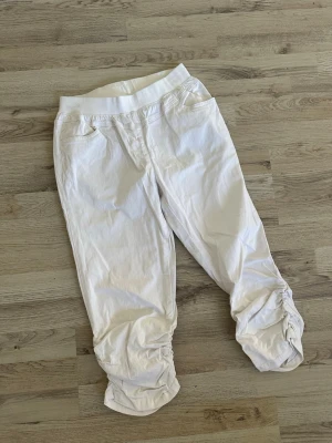 Vita leggings med rynkade benslut - Säljer ett par vita leggings med bred resår i midjan och rynkade detaljer vid bensluten. De har fickor framtill och är tillverkade i ett stretchigt bomullsmaterial. Perfekta för en avslappnad och trendig look.