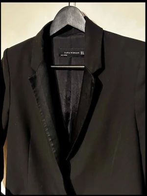 Zara Woman smokingkavaj med satinslag (Stl S) - Svart smokingkavaj från Zara Woman i stl S med fina satinslag framtill. Enkelknäppt modell med snygga ärmknappar. Utsidan är i absolut nyskick! Tvättlapparna på insidan är avklippta, annars inga anmärkningar. Se sista bilden för måttband! Mått: Bredd ca 44cm, längd ca 66cm, ärm ca 59cm. Perfekt trendigt basplagg till garderoben! Skriv vid frågor.