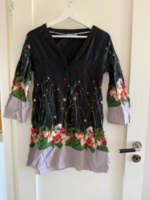 Blommig svart och grå klänning - Snygg korttunika/ klänning med svart överdel och grå nederdel, dekorerad med blommor. Klänningen har trekvartsärm och v-ringning med knappar framtill. Perfekt för dig som gillar unika mönster och sköna material.