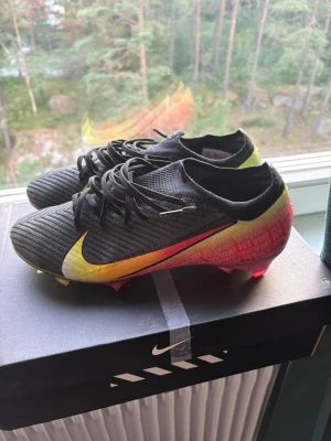 Nike Mercurial Vapor Vini Voa fly  - Säljer ett par Nike Mercurial Vapor fotbollsskor i svart med färgskiftande detaljer i gult, orange och rosa. Skorna har en låg profil, snörning och en ovandel i syntetmaterial för bra bollkontroll. Sulan har en cool gradient från gult till rosa och är designad för fart på planen., stl 43 passar även 42, skick 9/10, använd ett fåtal gånger
