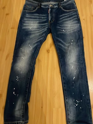 Mörkblå Dsquared2 jeans med stänk - Säljer ett par mörkblå jeans från Dsquared2 med coola vita färgstänk och slitningar. Jeansen har klassisk femficksmodell, raka ben och tydliga tvättade detaljer. Materialet är denim i bomull och bakfickan har D2-logga. Perfekta för en trendig streetwear-look.