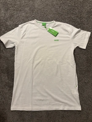 Vit t-shirt från Hugo Boss - Vit t-shirt från Hugo Boss med grönt BOSS-tryck på bröstet och axeln. Klassisk rund hals och korta ärmar. Tröjan är aldrig använd och har fortfarande lappen kvar från köp. Skriv gärna vid frågor och funderingar!😃✅