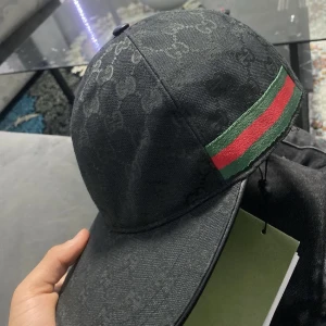 Svart Gucci keps med GG-mönster - Säljer en svart keps från Gucci med klassiskt GG-monogram över hela kepsen. På sidan finns den ikoniska gröna och röda randen. Kepsen har böjd skärm och justerbar passform. Perfekt för dig som vill ha en stilren accessoar med lyxig känsla. Taggen fins inte längre kvar!
