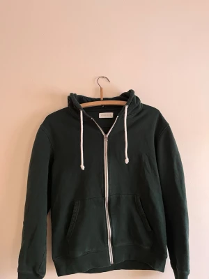 Mörkgrön hoodie med dragkedja Pier One - Mörkgrön hoodie från Pier One med vit dragkedja och vita dragsnören. Klassisk modell med huva och två fickor framtill. Tillverkad i mjukt bomullsmaterial som känns skönt mot huden. Perfekt för en avslappnad och stilren look.