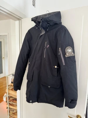 Svart parkas från Peak Performance - Snygg svart parkas från Peak Performance med flera praktiska fickor, dragkedja och tryckknappar framtill. Jackan har en hög krage och huva, samt en tydlig logotyp på ärmen. Perfekt för kalla dagar och riktigt stilren look. Small men är lätt fungerande för medium eller till och med slim Large. Kostade 9995kr på stadium 