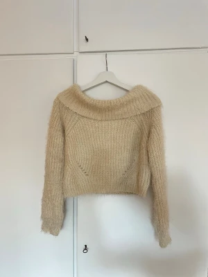 Beige fluffig bandeau tröja - Säljer en beige stickad bandeau tröja med fluffig känsla och bred krage. Tröjan har lång ärm och är något croppad i modellen. Perfekt för dig som gillar en mysig och trendig look. Väldigt mjuk och skön!