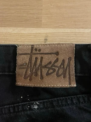 Svarta baggy  jeans från Stüssy - Säljer ett par svarta jeans från Stüssy med bred passform och raka ben. Jeansen har klassisk femficksdesign, knappgylf och ett läderpatch med Stüssy-loggan bak i midjan. Materialet är kraftig denim i bomull, perfekta för en streetwear-look.