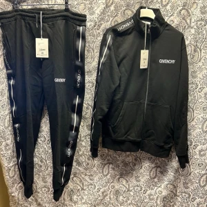 Svart tracksuit från Givenchy - Säljer ett svart tracksuit-set från Givenchy med vita detaljer längs ärmar och ben. Setet består av en zip-jacka med hög krage och två fickor samt matchande byxor med resår i midjan och muddar vid benslut. Givenchy-logga syns tydligt på både jacka och byxor.