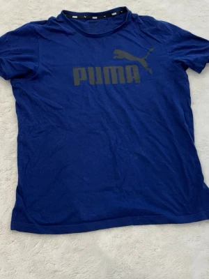 Blå Puma t-shirt med logga - Snygg blå t-shirt från Puma med stor svart logga och text på bröstet. Klassisk rund halsringning och korta ärmar. Tillverkad i mjuk bomull som känns skön mot huden. Perfekt för en avslappnad och sportig stil.
