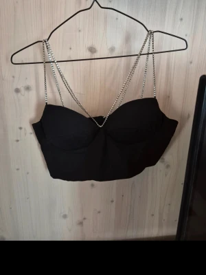 Svart topp med kedjeband - Snygg svart topp med tunna kedjeband som axelband och dragkedja i ryggen. Toppen har en croppad passform och vadderade kupor för extra stöd. Perfekt för att lyfta din outfit med en edgy touch.