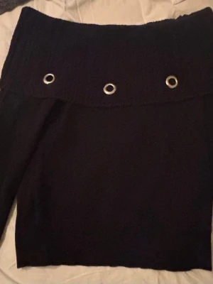 Svart off shoulder stickad tröja - Trendig svart stickad off shoulder-tröja med bred krage och fyra silvriga metallöljetter framtill och baktill. Figurnära passform och långa ärmar. Perfekt för dig som vill ha en unik och edgy look.