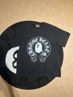 Svart Chrome Hearts x Bape T-shirt - Svart t-shirt med stor vit Chrome Hearts x Bape-logga på bröstet. Klassisk rund halsringning och korta ärmar. T-shirten har en streetwear vibe och är tillverkad i mjuk bomull för skön passform.
