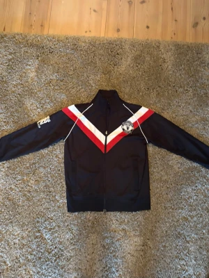 Manchester United Retro Track Jacket 06/07 - Snygg retro sport/trackjacka från Nike i svart med vita och röda V-formade detaljer över bröstet. Jackan har hel dragkedja, hög krage och logga på vänster bröst samt tryck på ena ärmen. Perfekt till träning eller streetwear. Storlek M passar 175-185. Äkta från  Classic Football Shirts