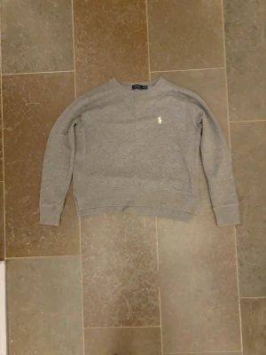 Grå sweatshirt från Polo Ralph Lauren - Klassisk grå sweatshirt från Polo Ralph Lauren med rund halsringning och den ikoniska vita broderade loggan på bröstet. Tröjan har ribbade muddar vid ärmslut och nederkant, perfekt för en avslappnad och stilren look.