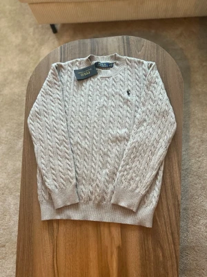 Beige kabelstickad tröja Polo Ralph Lauren - Beige kabelstickad tröja från Polo Ralph Lauren.  • Storlek: M • Passform: Herr / Unisex • Skick: Oanvänd – nyskick, med originaltag kvar • Färg: Beige • Detaljer: Klassisk kabelstickning, rund halsringning, ribbade muddar vid ärmslut och nederkant samt svart broderad logga på bröstet  Tröjan är helt oanvänd och i perfekt skick. Tidlös och stilren modell som passar både till vardag och mer uppklädda tillfällen.  Nypris: ca 1 700–1 900 kr