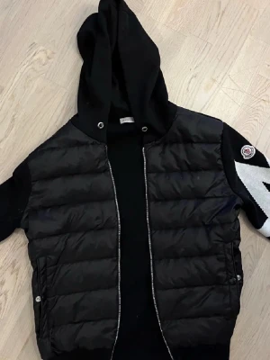 Moncler cardigan - Snygg svart moncler cardigan i storlek M med quiltad front och stickade ärmar. Jackan har dragkedja, huva med snörning och ett stor vitt M på ärmen samt Moncler logga. Perfekt för dig som vill ha en skön och trendig look. Svarar alla meddelanden direkt och fraktar alltid samma dag som köp:)