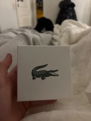 Silverfärgat armband från Lacoste - Säljer ett stilrent silver armband från Lacoste med klassiskt silverfärgad metall. Armbandet levereras i originalförpackning med den ikoniska krokodilloggan. Perfekt accessoar för dig som gillar en clean och modern look.
