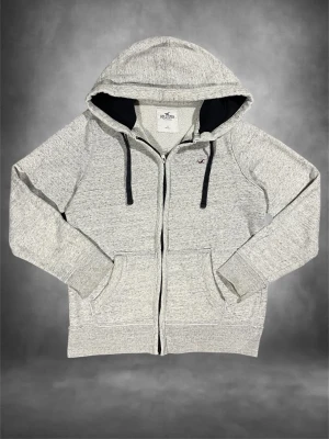 Ljusgrå zip hoodie från Hollister - Ljusgrå hoodie från Hollister med dragkedja, svart fodrad huva och svarta snören. Klassisk logga på bröstet och två stora fickor framtill. Tillverkad i mjukt bomullsmaterial som är perfekt för chill dagar.