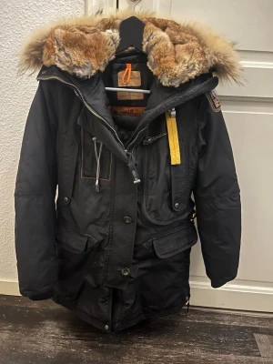 Svart Parajumpers parkas med pälskrage - Säljer en svart parkas från Parajumpers med stor pälshuva och  ikonisk gul rem vid halsen. Jackan har flera fickor, grov dragkedja och patch på ärmen. Perfekt för kalla dagar och riktigt snygg streetstil. Lite slitet på skinndetaljerna, därför bra pris. Storlek S