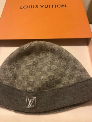 Grå Louis Vuitton mössa - Snygg grå mössa från Louis Vuitton med klassiskt rutmönster och LV-logga framtill. Mössan är stickad och har en vikt kant. Perfekt accessoar för att lyfta din stil under kalla dagar.