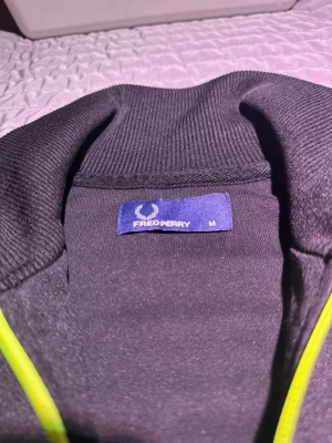 Svart Fred Perry ziptröja med neonränder - Säljer en svart ziptröja från Fred Perry med neonfärgade gula ränder längs ärmarna och dragkedjan. Tröjan har hög krage, ribbade muddar och Fred Perry-logga broderad på bröstet. Perfekt för en sportig och trendig look. Färgvariant som inte längre finns i butik och är svår att hitta idag.
