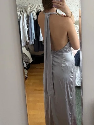 Ljusblå/silver långklänning med slits och öppen rygg - Snygg ljusblå/silverfärgad långklänning i satinliknande material med halterneck och öppen rygg. Klänningen har en knytning i nacken och en slits framtill. Jättefin till balen mm. 