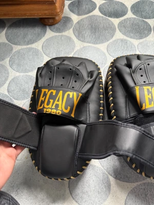 Legacy 1980 svart mitts set - Säljer ett par svarta mittsar från Legacy 1980, perfekta för boxning eller thaiboxning. Tillverkade i slitstarkt syntetläder med gul logga och detaljer. Justerbart kardborreband för bra passform och extra vaddering för skydd. Handgjorda i Thailand.