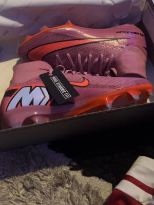Nike Mercurial rosa fotbollsskor elite - Säljer ett par Nike Mercurial fotbollsskor i en snygg rosa färg med orange och svart detaljering samt stor M-logga på hälen. Skorna har högt skaft, snörning och en slitstark yttersula med dobbar för bra grepp på planen. Materialet är syntetisk. Testa dom men var för små, använd endast 1 träning.