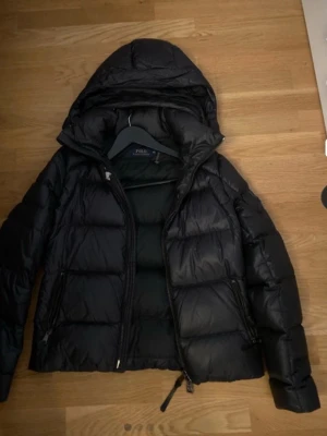 Svart pufferjacka från Polo Ralph Lauren - Snygg svart pufferjacka från Polo Ralph Lauren med huva och dragkedja framtill. Jackan har en klassisk quiltad design, två sidofickor med dragkedja och är perfekt för kyliga dagar. Materialet är glansigt och ger en modern look.