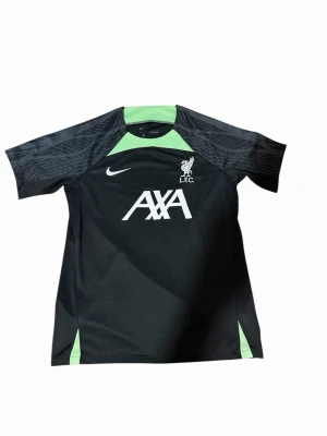 Nike Liverpool träningströja svart/grön - Snygg svart träningströja från Nike med Liverpool FC-logga och AXA-tryck på bröstet. Tröjan har gröna detaljer vid hals och sidor samt grafiskt mönster på axlarna. Tillverkad i lätt och funktionellt material, perfekt för träning.