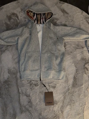 Grå hoodie från Burberry med dragkedja - Snygg grå hoodie från Burberry med dragkedja framtill och klassiskt rutigt foder i huvan. Tröjan har fickor på sidorna och justerbar dragsko i huvan. Perfekt för en avslappnad och trendig look. Materialet känns mjukt och bekvämt. 