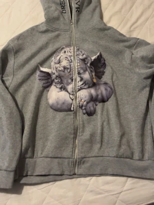 Grå hoodie med ängeltryck från Reality - Säljer en grå zip hoodie från Reality med stor grafisk ängel på framsidan och texten 'REALITY' broderad på ryggen. Hoodien har huva och dragkedja hela vägen. Perfekt för dig som gillar streetwear och unika prints.