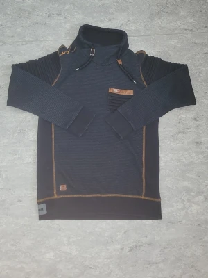 Mörkblå hoodie med bruna detaljer - Snygg mörkblå hoodie från wam denim med bruna kontrastsömmar och detaljer. Hoodien har en hög krage med dragsko, ribbade ärmar och en unik ficka på bröstet med brunt kantband och en liten flaggdetalj. Materialet är mjukt och bekvämt, perfekt för en avslappnad stil.