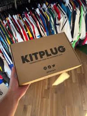 Kitplug Mystery Football Shirt Box - Mystery box från Kitplug fylld med fotbollströjor i olika färger och mönster från kända lag. Perfekt för dig som vill överraska dig själv med unika matchtröjor. Kartongen är brun med svart text och innehållet varierar mellan olika lag och stilar.
