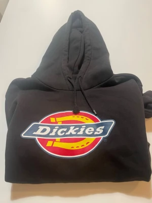 Svart Dickies hoodie med logga - Säljer en svart hoodie från Dickies med stor färgglad logga på bröstet. Klassisk huva med dragsko och mjukt material som känns skönt mot huden. Perfekt för dig som gillar streetwear och vill ha en enkel men snygg look.