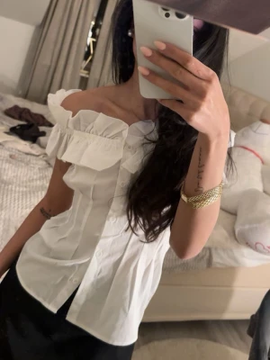 Vit offshoulder blus med volang - Superfin vit offshoulder blus med volangdetaljer längs axlarna och knappar framtill. Blusen har en figurnära passform och är tillverkad i ett lätt tyg som känns lufftigt 