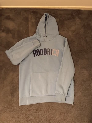 Ljusblå HOODRICH hoodie med huva - Snygg ljusblå hoodie från HOODRICH med stor huva och magficka. Framsidan har HOODRICH-loggan i svart och silver. Tillverkad i mjukt material, perfekt för chill dagar. Klassisk passform och ribbade muddar vid ärmar och nederkant.