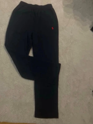 Mörkblå mjukisbyxor från Ralph Lauren - Säljer ett par svarta mjukisbyxor från Ralph Lauren med den klassiska röda loggan broderad på vänster sida. Byxorna har resår i midjan och raka ben, perfekta för chill dagar. Materialet känns mjukt och bekvämt.