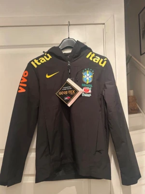 Svart Brasil Nike vindjacka Gore-Tex - Snygg svart vindjacka från Nike med Brasil-logga, gula och orangea detaljer samt sponsorer som Itaú och Vivo. Jackan har huva, dragkedja och är tillverkad i slitstarkt Gore-Tex-material som står emot regn och vind. Perfekt för dig som gillar sportig stil.