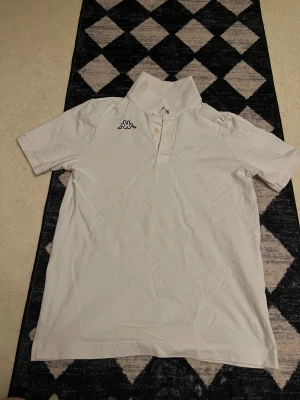 Vit pikétröja från Kappa - Säljer en klassisk vit pikétröja från Kappa med korta ärmar och krage. Tröjan har knappar framtill och Kappa-logga broderad på bröstet samt tryckt på kragen. Perfekt till en sportig och avslappnad stil.