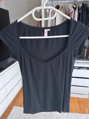 Svart topp med spetskant från Nelly - En svart, enkel topp från H&M med korta ärmar och en fin spetskant runt den breda, rundade urringningen. Tunn och mjuk i materialet, perfekt att styla till olika looks. Klassisk och stilren design.