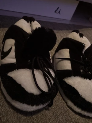 Svarta och vita fluffiga tofflor Nike  - Mysiga tofflor i svart och vitt med fluffig yta och snörning framtill. De har en sportig look med en tydlig logga på sidan och stora tofsar på ovansidan. Perfekta för att hålla fötterna varma hemma. Materialet är mjukt och plyschliknande.