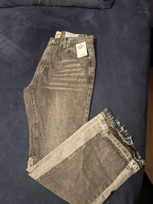 Gråa jeans med råa kanter Gallery Dept - Unika grå jeans från Gallery Dept med tvättad look och råa, fransiga kanter ovanför knäna. Jeansen har fem fickor, raka ben och cool patchwork-detalj på baksidan. Perfekta för dig som gillar streetwear och vill sticka ut.