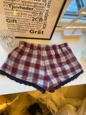 Rutiga pyjamasshorts med spetskant - Säljer ett par mysiga pyjamasshorts i rött, vitt och blått rutigt mönster. Shortsen har mörkblå spetskant nedtill och en söt rosett framtill i midjan. Perfekta för chillkvällar hemma. Mjukt och bekvämt material.🤍