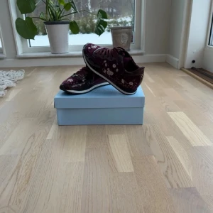 Vinröda Philippe Model sneakers - Snygga vinröda sneakers från Philippe Model med blommönster i vitt och rosa. Skorna har en låg siluett, vita och svarta detaljer på sulan samt glansig finish. Perfekta för dig som vill sticka ut med en unik och trendig look.
