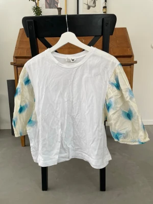 Vit topp med mönstrade ärmar Bimba Y Lola - Unik vit topp från Bimba Y Lola med halvtransparenta, puffiga ärmar i vitt med blått och turkost blommönster. Rak passform och rund halsringning. Perfekt för dig som vill sticka ut lite extra.