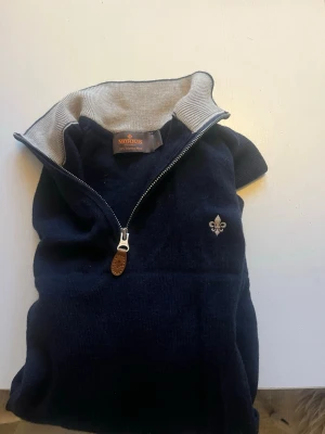 Mörkblå halvzip-tröja från Morris - Mörkblå stickad tröja från Morris med halv dragkedja och kontrastfärgad insida i kragen. Tröjan har en broderad lilja på bröstet och en snygg läderdetalj på dragkedjan. Perfekt för en stilren och avslappnad look.