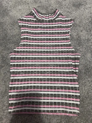 Randig ribbad ärmlös topp - Säljer en ärmlös topp med hög krage i ribbat material. Toppen har ett randigt mönster i svart, vitt och rosa och är croppad i modellen. Perfekt för dig som gillar en färgglad och trendig stil.