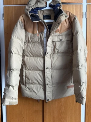 Beige dunjacka med läderdetaljer - Snygg beige dunjacka från Solid med bruna läderdetaljer på axlarna och huva med dragsko. Jackan har stora fickor med lock och tryckknapp samt quiltad design. Perfekt för kalla vinterdagar och riktigt varm.