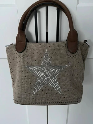 Beige handväska med stjärndetalj - Snygg beige handväska i canvas med bruna handtag i konstläder. Framsidan har en stor stjärna i silverfärgade nitar och små prickar för extra bling. Väskan har dragkedja baktill och metallfästen för axelrem. Perfekt för dig som gillar trendiga detaljer!